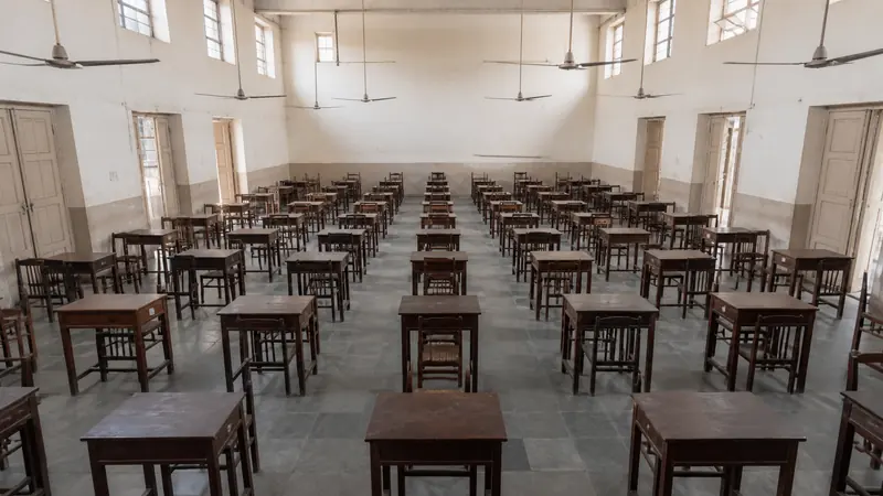 Widely spaced single desks arranged in an empty Indian school hall beneath ceiling fans, ready for an examination.