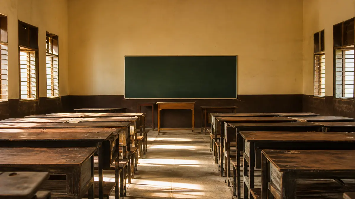 Rows of empty wooden benches and desks in a sunlit Indian school classroom with a blank blackboard.
