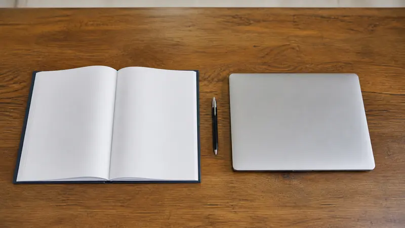 A bound fee ledger lying beside a closed laptop on a school accounts desk, two record-keeping eras side by side.