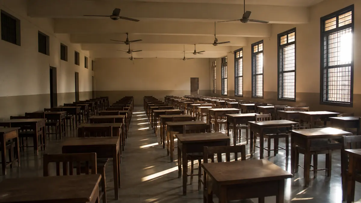 Rows of empty single wooden desks arranged for an examination in a sunlit Indian school hall, ceiling fans overhead.