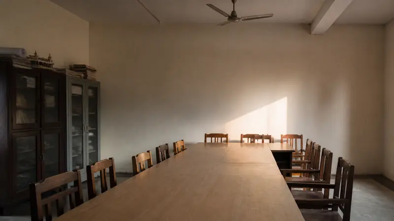 An empty school meeting room with chairs drawn up to a long table and afternoon light falling across a bare wall.